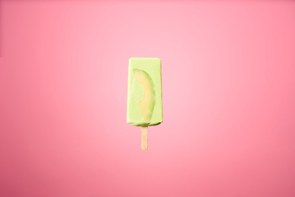 A green avocado popsicle with a slice of avocado is placed in a pink background