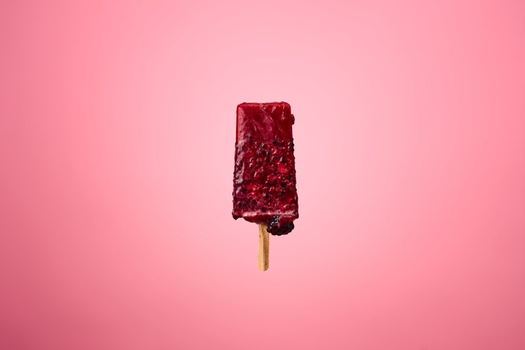 A dark red popsicle is placed in front of a pink background