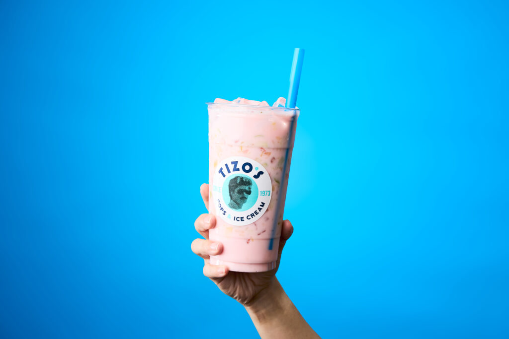 A cup of pink juice and fruit chunks with a large blue straw is being held in front of a blue background