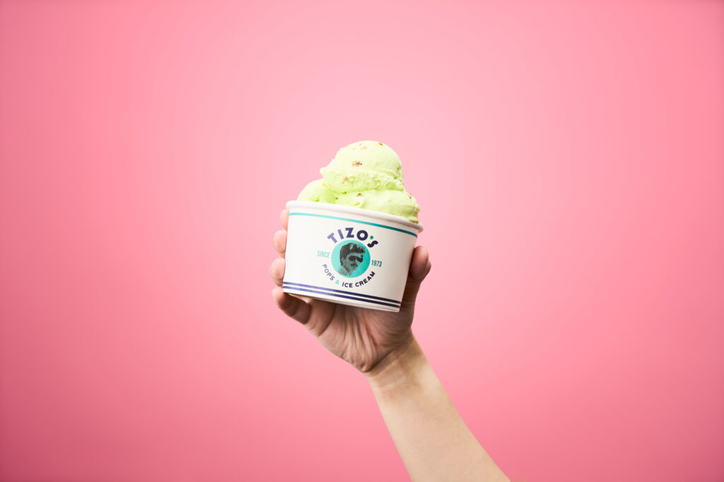 A scoop of pistachio ice cream inside a white cup is being held up in front of a pink background