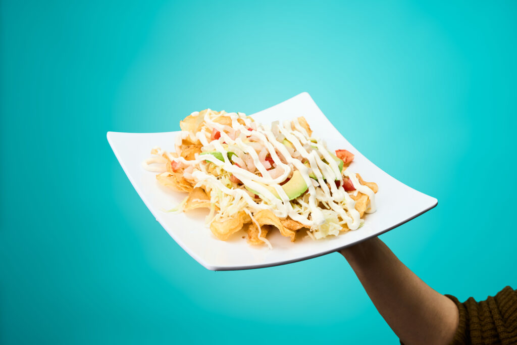 Plate of chicharron nachos topped with avocado slices being held up in front of teal background
